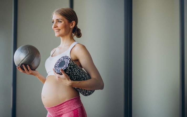 Pregnant woman wearing exercise gear and holding roll mat with exercise ball.