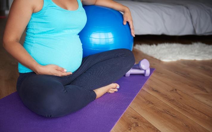 Woman holding pregnancy bump while exercising on purple yoga mat with blue exercise ball and small weights.